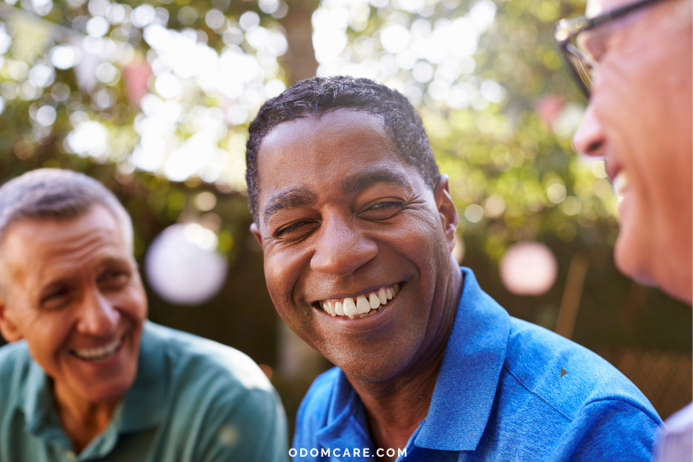 A close-up of three elderly men sitting together outdoors, smiling and laughing. The focus is on a man in a blue shirt in the center, with two other men on either side. The background is a blurred natural setting with greenery and soft lighting. The website 'ODOMCARE.COM' is displayed at the bottom.
